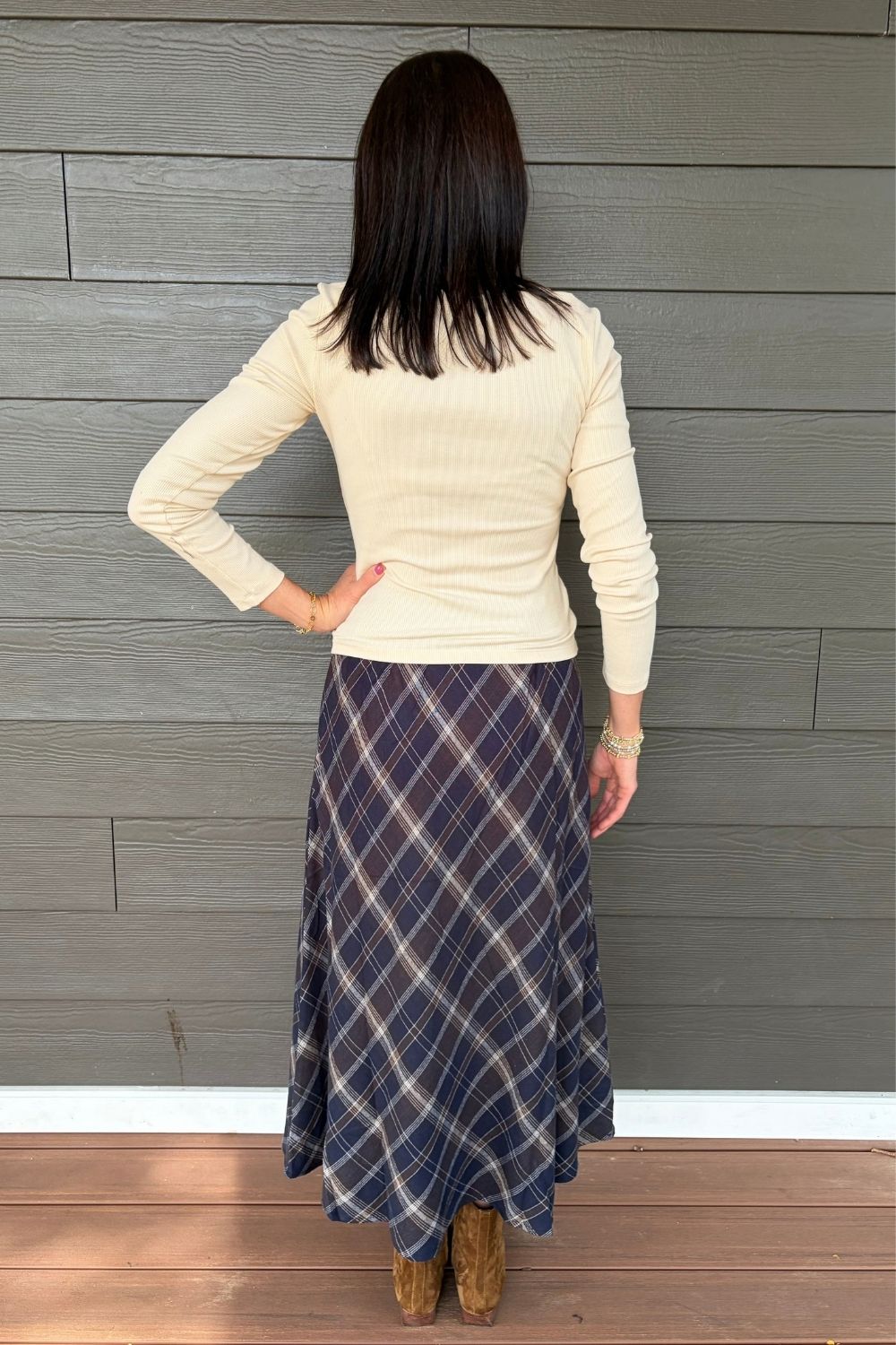 A woman shown from the back wearing a cream ribbed long-sleeve top and a navy blue plaid midi skirt, standing against a gray wood-paneled wall.