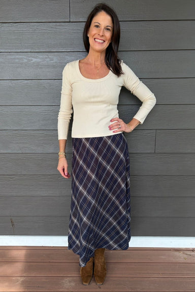 A woman stands against a gray wood-paneled wall wearing a cream ribbed long-sleeve scoop-neck top and a navy blue plaid maxi skirt, paired with brown suede boots.