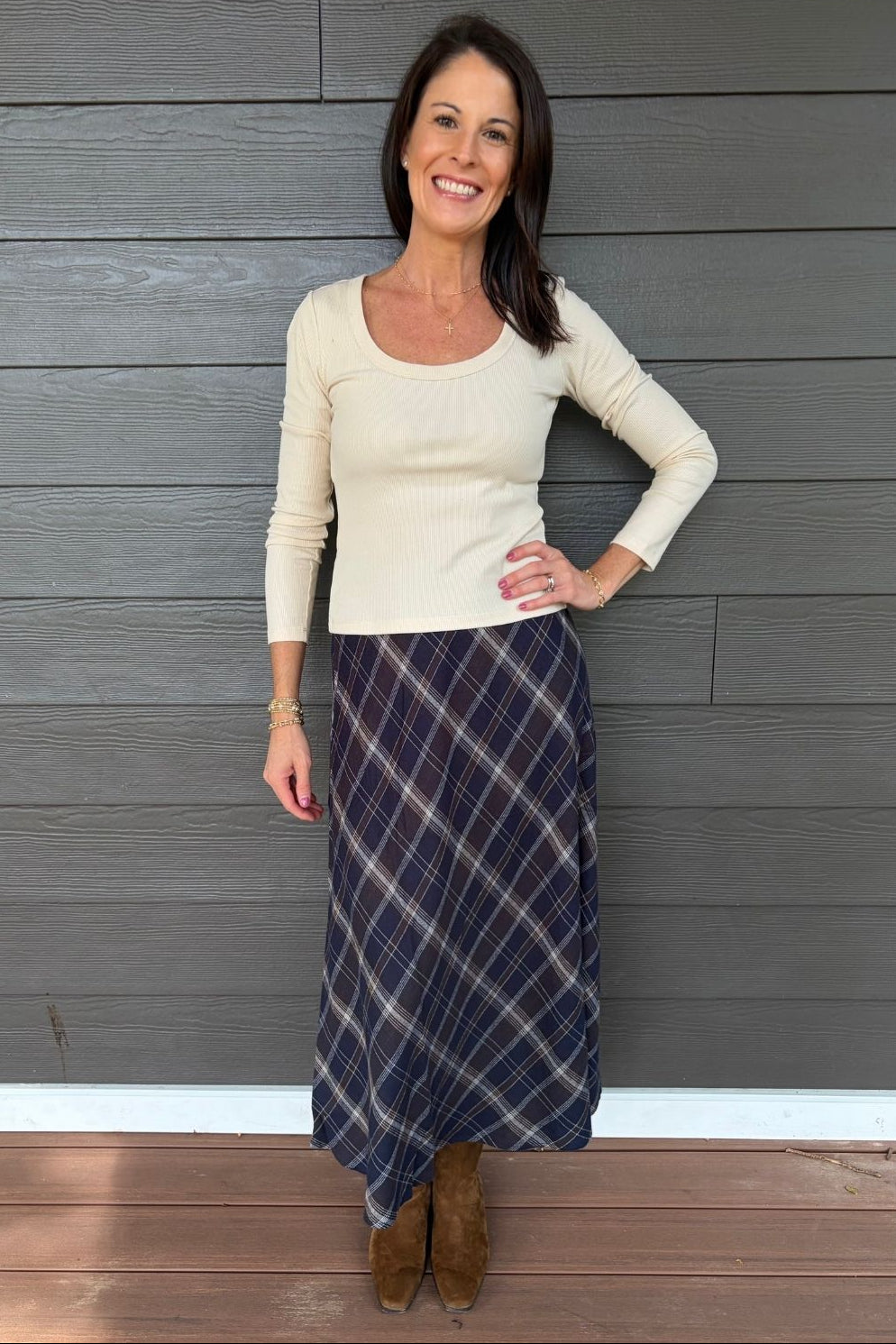 A woman stands against a gray wood-paneled wall wearing a cream ribbed long-sleeve scoop-neck top and a navy blue plaid maxi skirt, paired with brown suede boots.