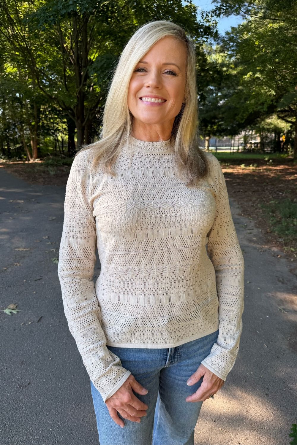 A smiling blonde woman wearing a cream-colored, textured knit mock-neck sweater and blue jeans, standing outdoors on a paved path with trees in the background.