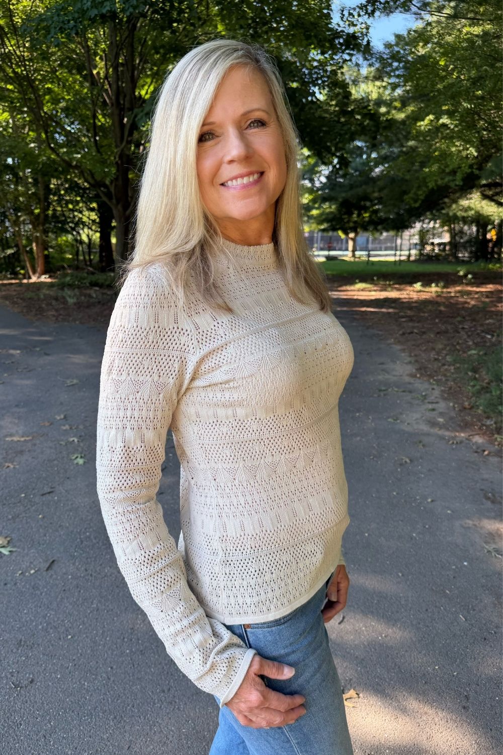A smiling blonde woman wearing a cream-colored, textured knit mock neck sweater and blue jeans, standing outdoors on a paved path surrounded by green trees.