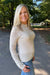 A smiling blonde woman wearing a cream-colored, textured knit mock neck sweater and blue jeans, standing outdoors on a paved path surrounded by green trees.