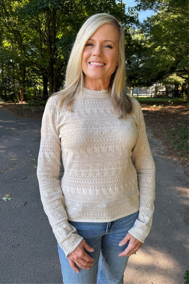 A smiling blonde woman wearing a cream-colored, textured knit mock-neck sweater and blue jeans, standing outdoors on a paved path with trees in the background.