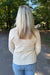 A woman viewed from behind wearing a cream-colored, long-sleeve knit sweater with textured geometric patterns, paired with blue jeans, standing on an outdoor paved path surrounded by green trees.