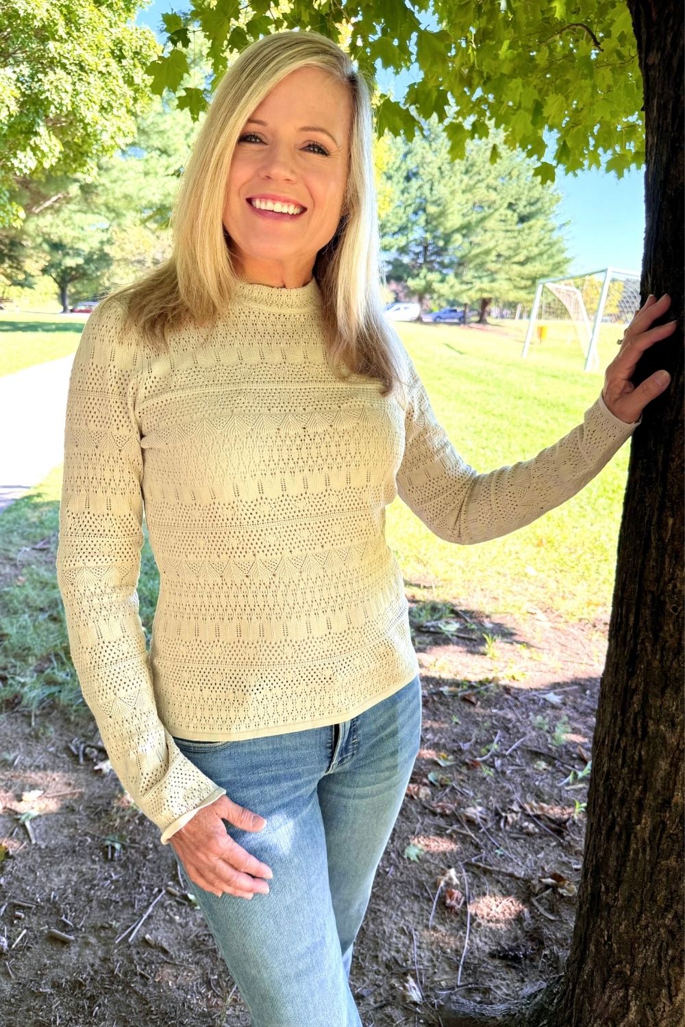 A smiling blonde woman wearing a cream-colored, long-sleeve knit mock neck sweater and blue jeans, standing outdoors next to a tree in a park.
