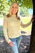 A smiling blonde woman wearing a cream-colored, long-sleeve knit mock neck sweater and blue jeans, standing outdoors next to a tree in a park.