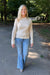 A woman walking on a paved path in a park, wearing a cream-colored textured knit sweater and blue flared jeans.