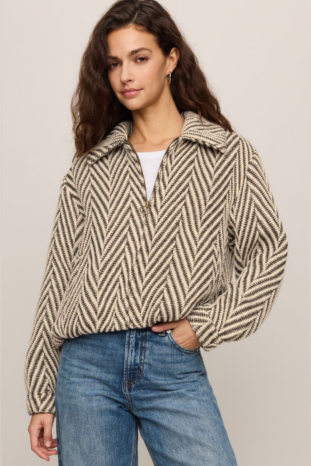 A woman wearing a beige and black herringbone patterned quarter-zip pullover sweater with a half-collar, styled with a white top and blue jeans against a neutral studio background.