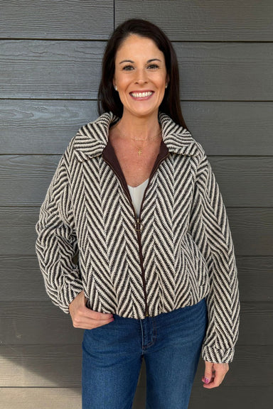 A woman smiling and wearing a zip-up jacket with a black and white chevron pattern, brown trim, and a brown zipper, paired with blue jeans, standing against a dark gray wood-paneled wall.
