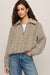 A woman wearing a beige and black herringbone patterned quarter-zip pullover sweater with a half-collar, styled with a white top and blue jeans against a neutral studio background.