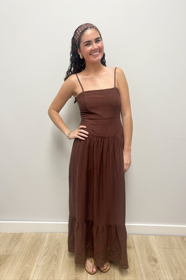 A woman modeling a brown spaghetti-strap maxi dress with a ruched bodice and tiered skirt, standing against a plain light gray wall on a wood floor.