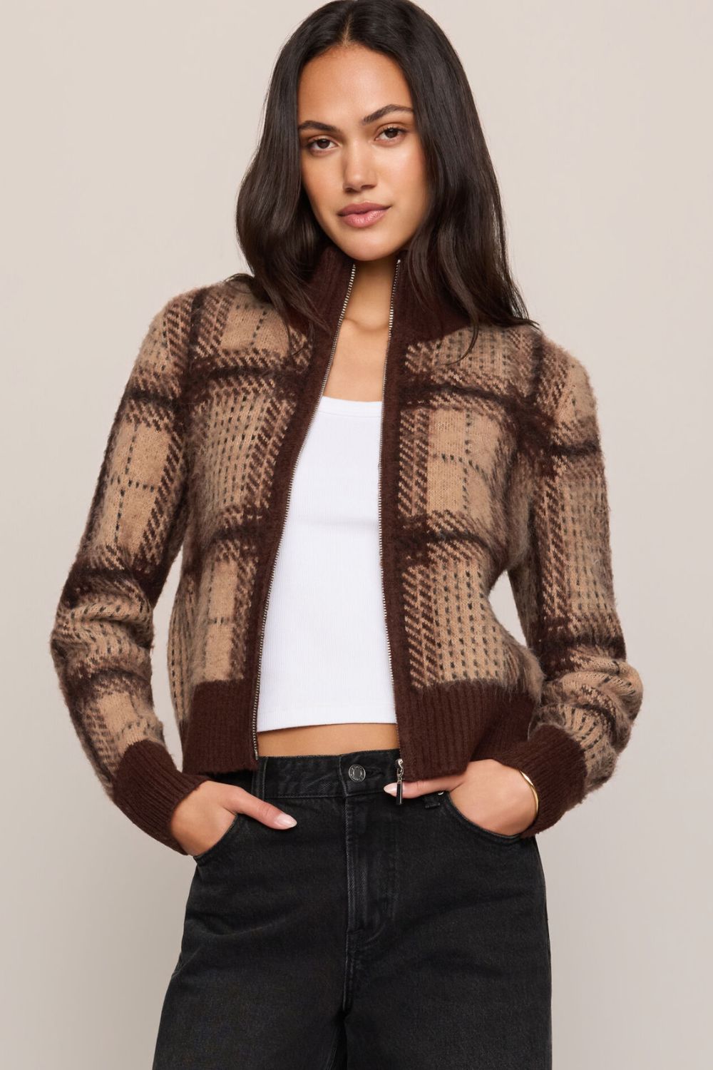A woman modeling a brown and beige plaid knit cardigan with a mock neck and ribbed cuffs, worn over a white crop top and black jeans against a solid light beige background.