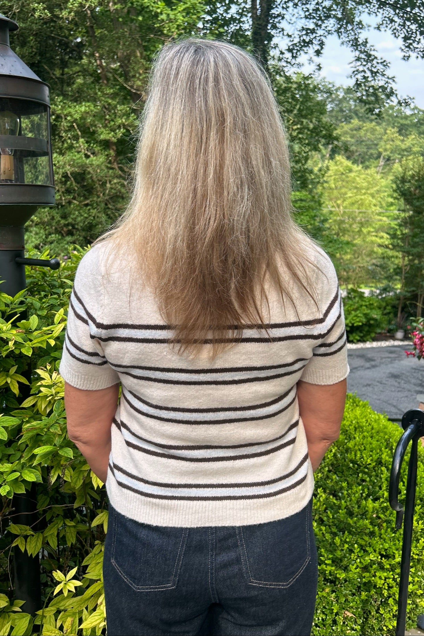 A woman viewed from behind wearing a cream knit short-sleeve top with thin black horizontal stripes and dark blue jeans, standing outdoors in front of green bushes and trees.
