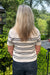 A woman viewed from behind wearing a cream knit short-sleeve top with thin black horizontal stripes and dark blue jeans, standing outdoors in front of green bushes and trees.