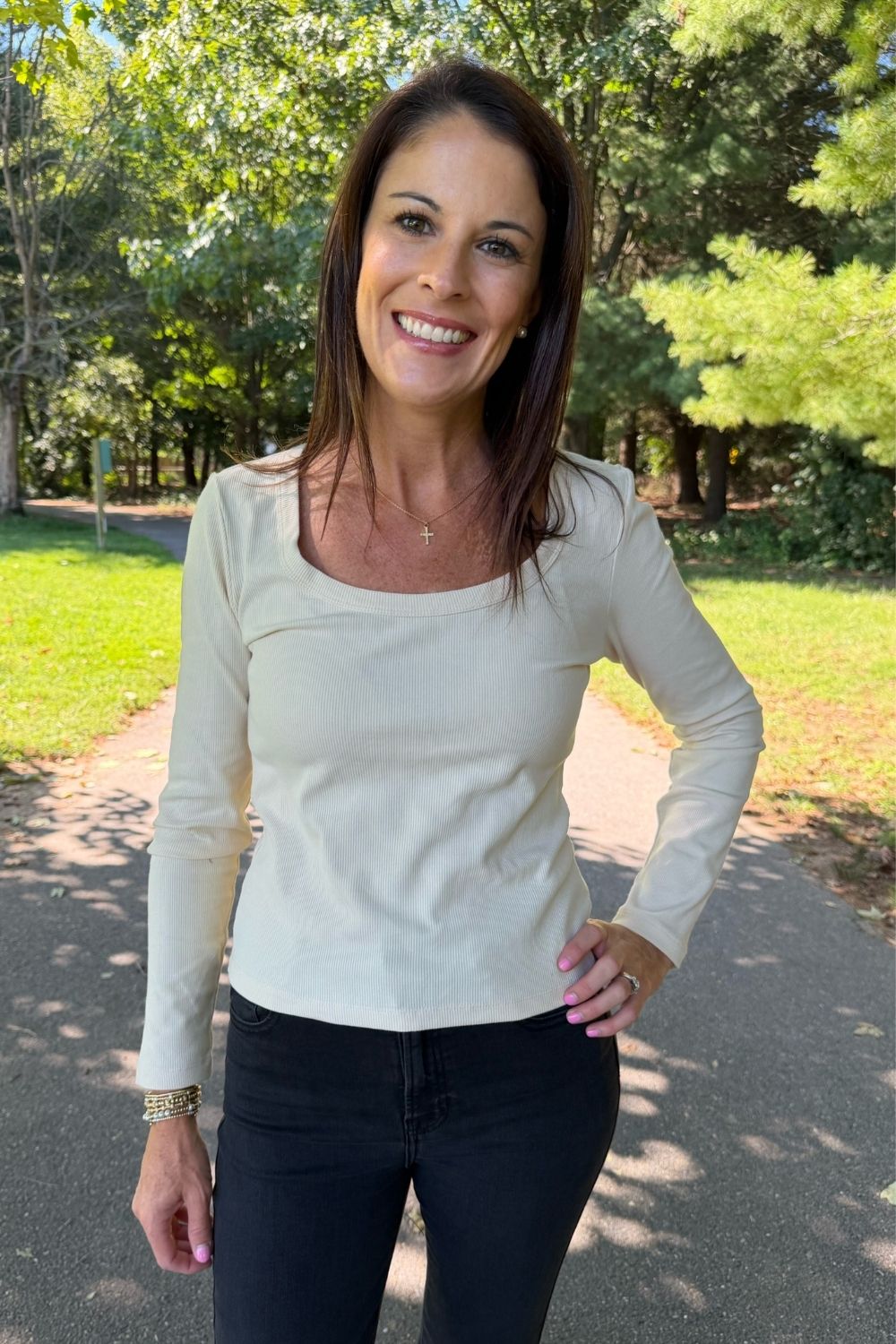A woman smiling outdoors on a paved path wearing a fitted long-sleeve scoop-neck top in a light cream color, paired with black pants.