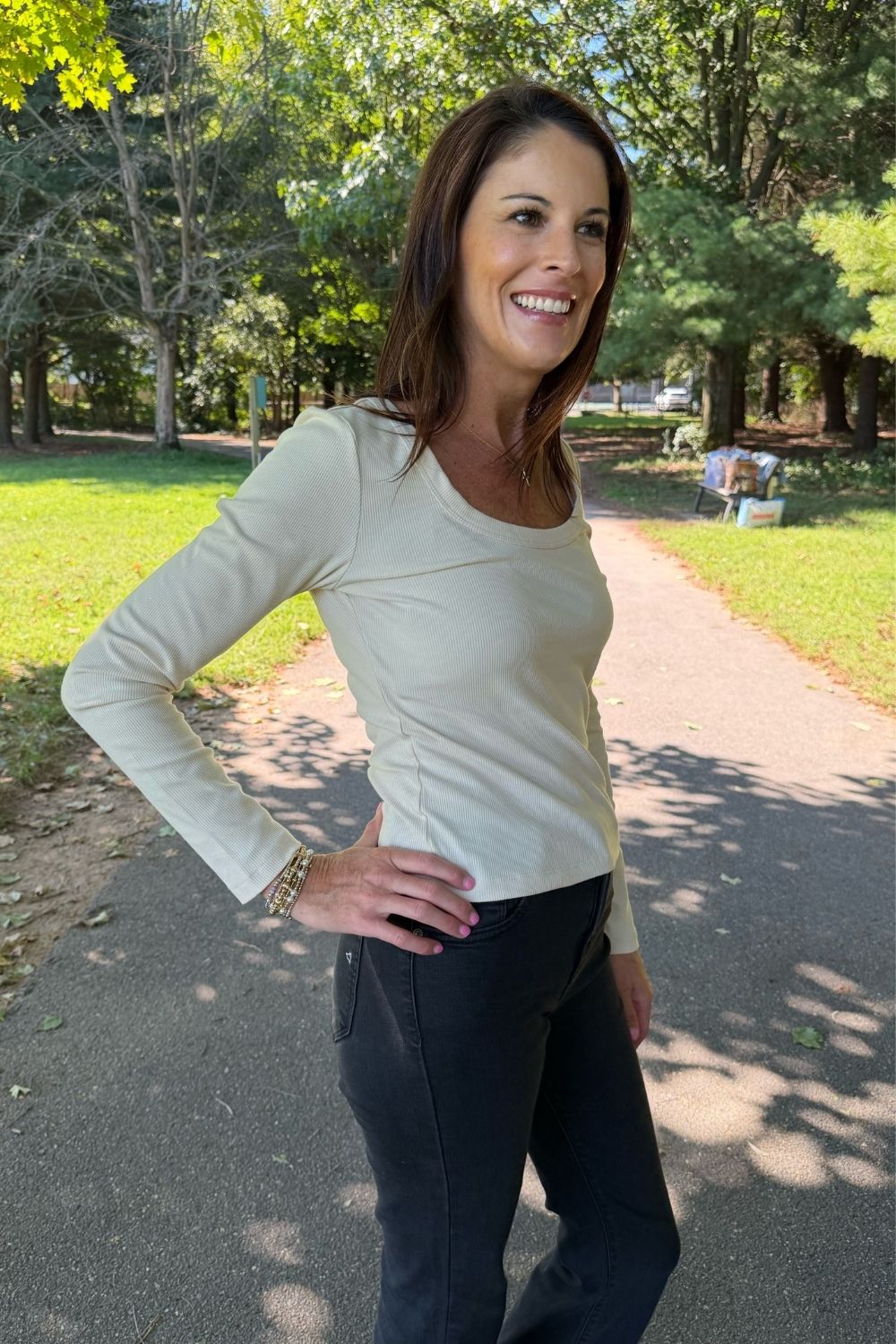 A woman smiling outdoors on a park path, wearing a fitted long-sleeve scoop-neck top in a light cream or off-white color, paired with dark pants.
