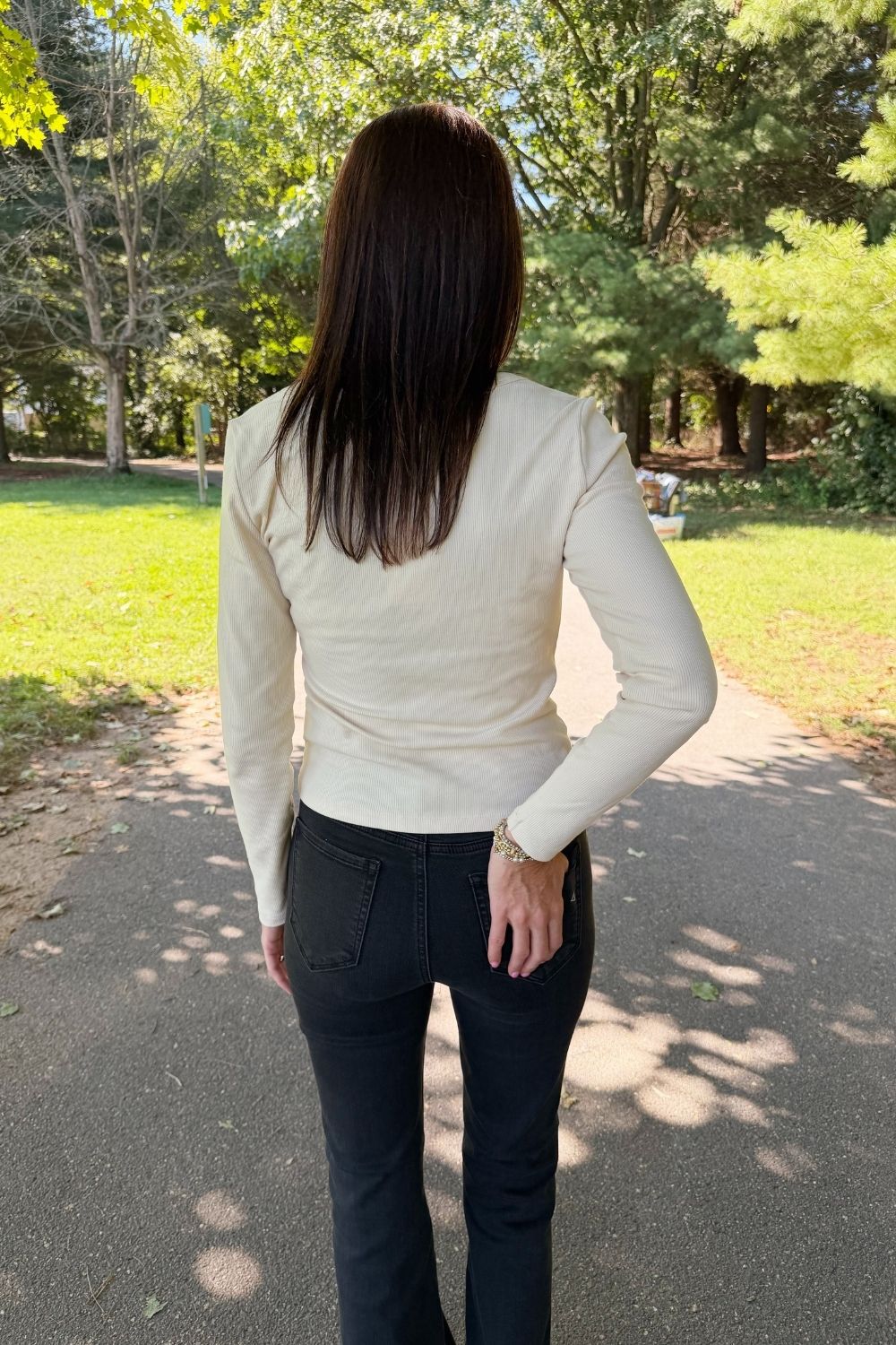 A woman viewed from behind wearing a fitted cream ribbed long-sleeve top and dark charcoal pants, standing on a paved path in a sunny park with trees and grass in the background.