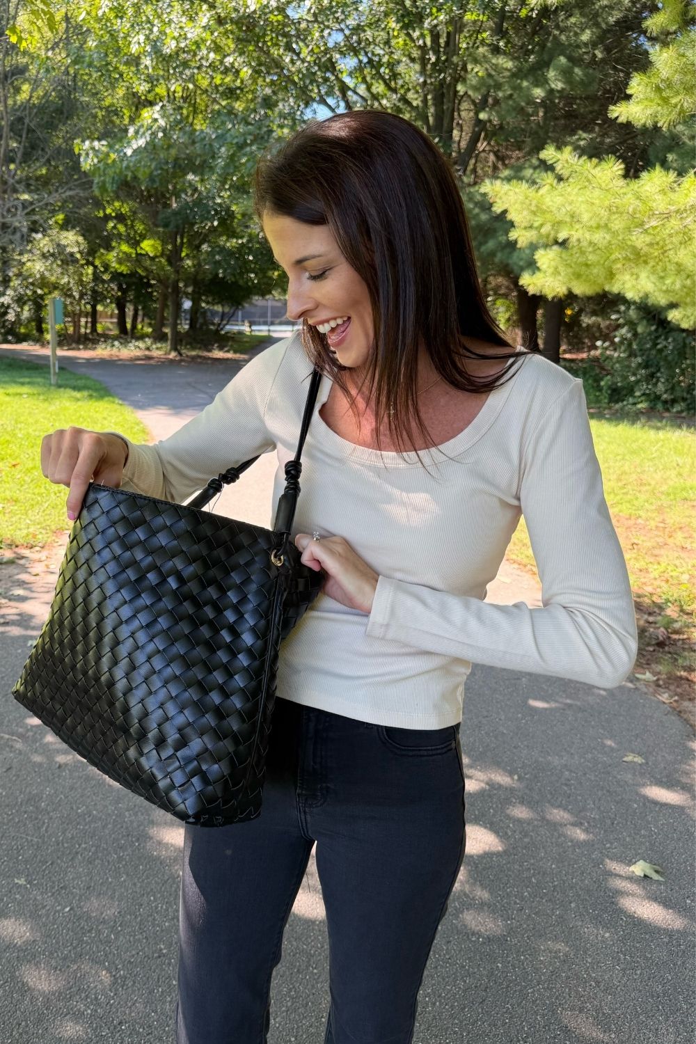 A woman outdoors on a paved path carrying a large black woven tote bag over her shoulder. She is wearing a cream long-sleeve top and dark pants, with trees and grass in the background.
