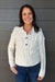 A smiling woman with dark hair stands against a gray wood-plank wall wearing a cream cable-knit cardigan with a buttoned collar and dark buttons, paired with blue jeans.