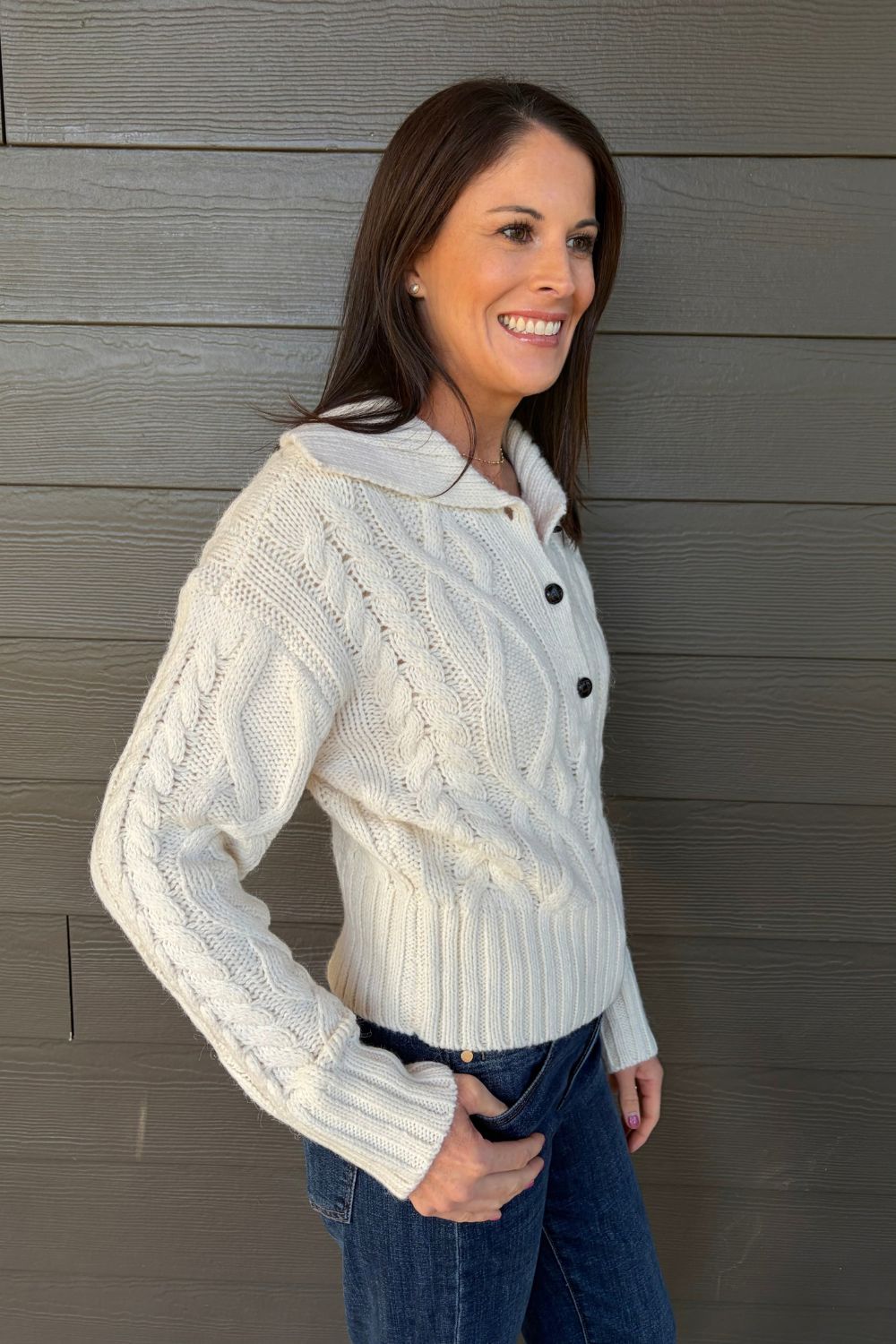 A woman wearing a cream cable-knit button-front cardigan with a collar, paired with blue jeans, standing against a gray wood-paneled wall.