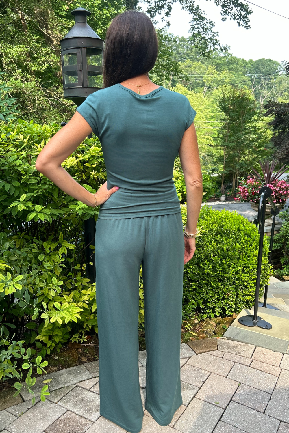 A woman viewed from behind wearing a sage green short-sleeve top and matching wide-leg pants, standing on a stone patio in a garden setting.