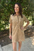 A woman wearing a tan corduroy shirt dress with short sleeves, a button-down front, and two chest pockets, standing outdoors in front of green bushes.