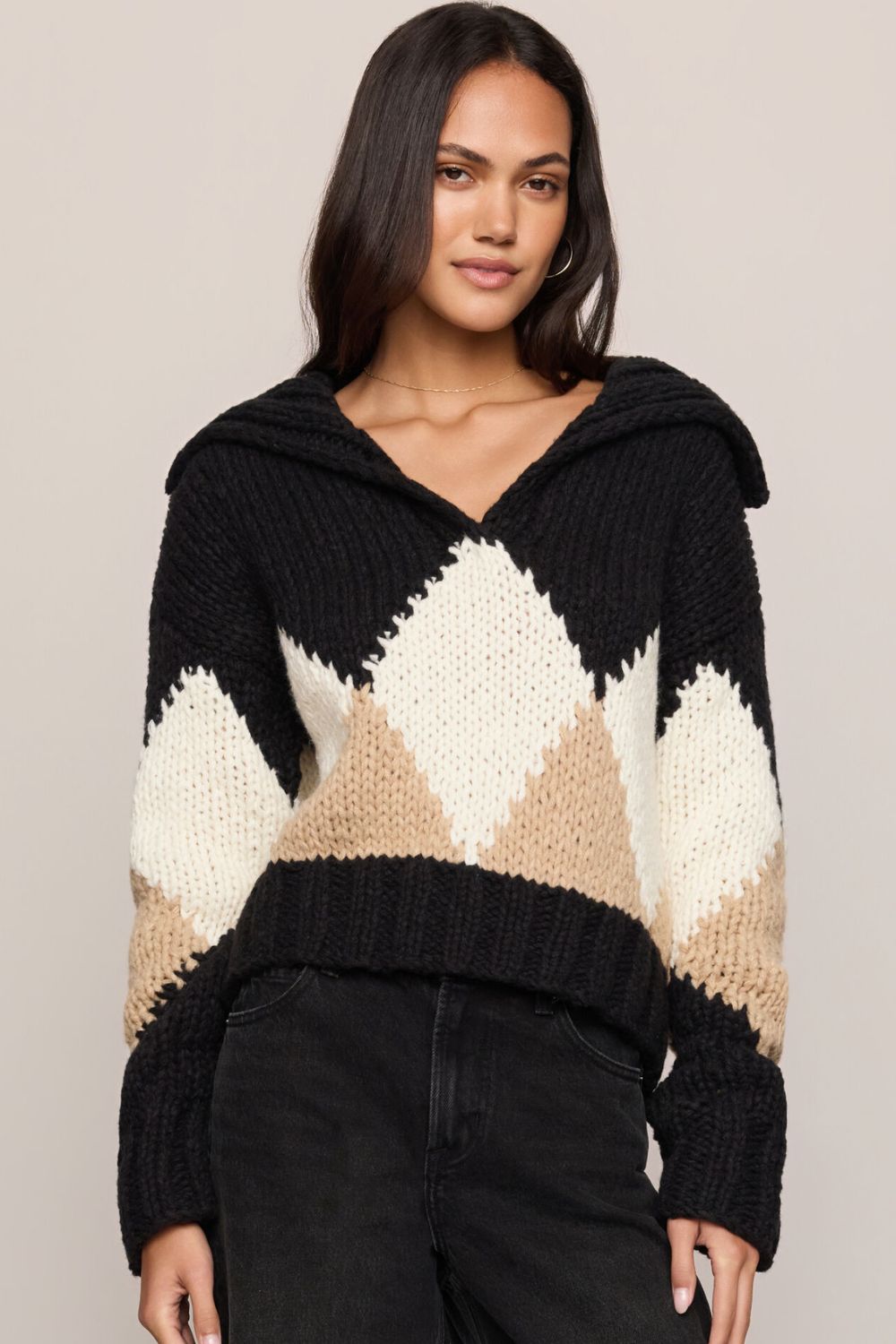 A woman wearing a chunky knit hooded sweater with a black, cream, and tan diamond pattern, paired with black jeans, against a plain light gray studio background.