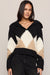 A woman wearing a chunky knit hooded sweater with a black, cream, and tan diamond pattern, paired with black jeans, against a plain light gray studio background.