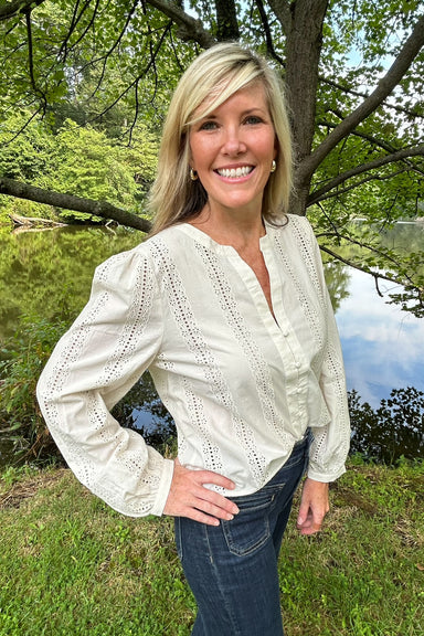 A smiling blonde woman wearing a cream eyelet long-sleeve blouse and dark blue jeans, standing outdoors in front of a tree and grassy area near water.