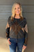 A smiling blonde woman wearing a cropped argyle cardigan in dark gray, brown, and beige with blue jeans, standing against a gray wood-paneled wall.