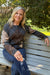 A woman with blonde hair sits on a gray wooden park bench outdoors, wearing a dark gray and brown argyle knit cardigan with blue jeans.
