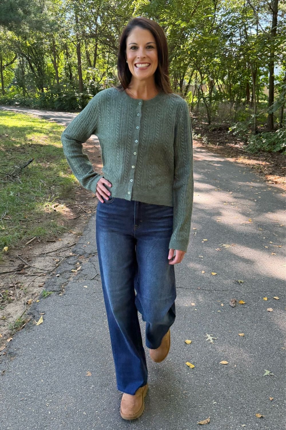 A woman stands on a paved path in a wooded outdoor setting, wearing a sage green cable-knit cardigan with buttons, blue jeans, and tan boots.
