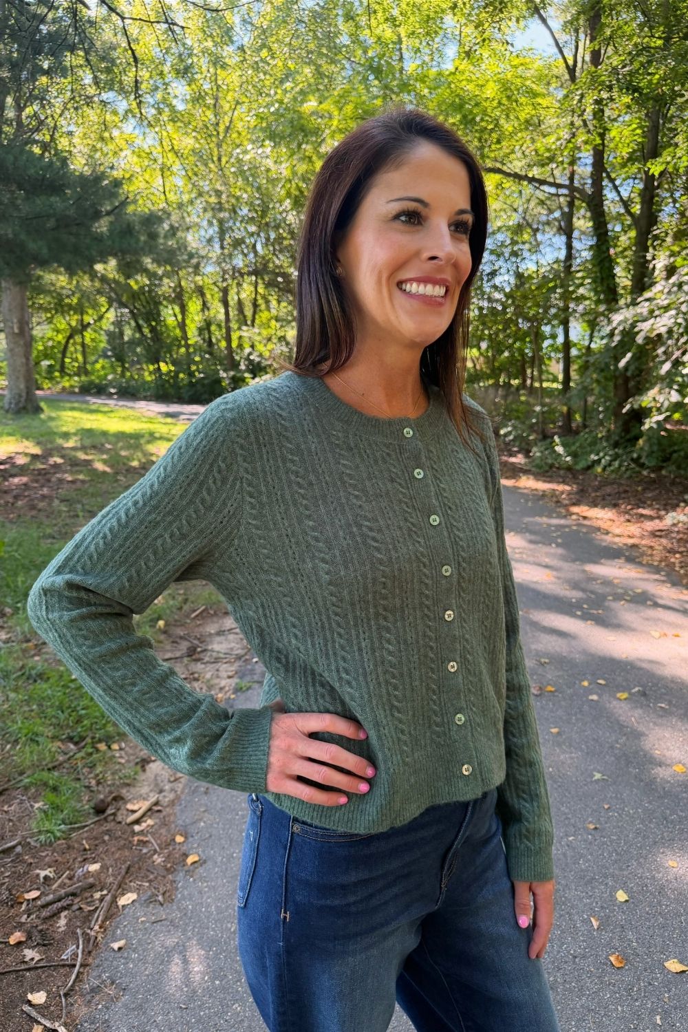 A woman wearing a sage green cable-knit button-up cardigan sweater and blue jeans, standing outdoors on a paved path surrounded by trees.