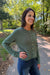 A woman wearing a sage green cable-knit button-up cardigan sweater and blue jeans, standing outdoors on a paved path surrounded by trees.