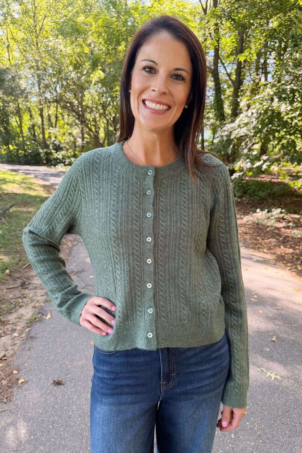 A woman wearing a sage green cable-knit button-up cardigan and blue jeans, standing outdoors on a paved path with trees in the background.