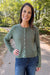 A woman wearing a sage green cable-knit button-up cardigan and blue jeans, standing outdoors on a paved path with trees in the background.