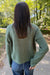 A person viewed from behind wearing an olive green cable-knit sweater and blue jeans, standing on a paved path in a wooded outdoor setting.