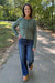 A woman stands on a paved path in a wooded outdoor setting, wearing a sage green cable-knit cardigan with buttons, blue jeans, and tan boots.