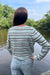 A woman viewed from behind wearing a light sage green knit sweater with dark brown and white horizontal stripes, standing by a river with trees in the background.