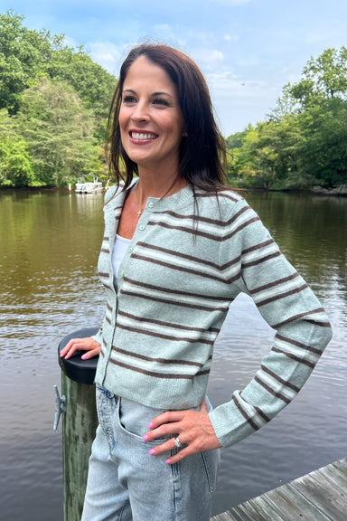 A woman smiling outdoors by a lake, wearing a light sage green and brown striped knit cardigan over a white top and light blue jeans.