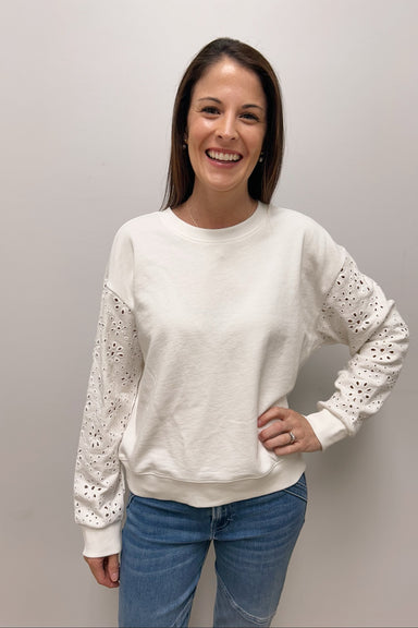 Woman wearing a white sweater with lace details and blue jeans against a plain background