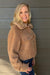 A smiling blonde woman wearing a cropped tan faux-shearling jacket with a wide collar and front flap pocket, paired with blue jeans, standing against a dark gray wood-paneled wall.