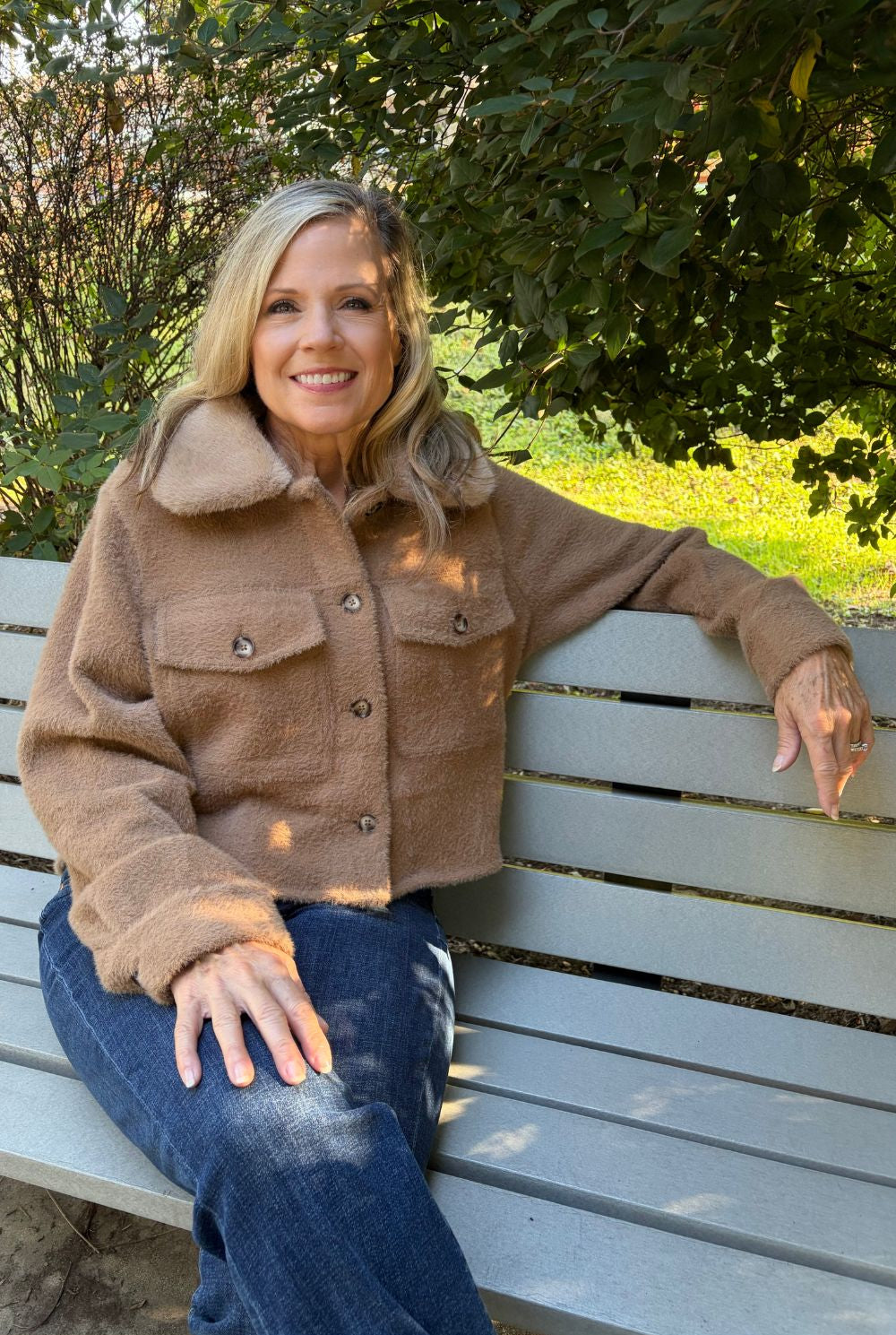 A smiling blonde woman sits on a gray wooden park bench outdoors, wearing a cropped brown teddy fleece jacket with a large collar and blue jeans.