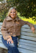 A smiling blonde woman sits on a gray wooden park bench outdoors, wearing a cropped brown teddy fleece jacket with a large collar and blue jeans.