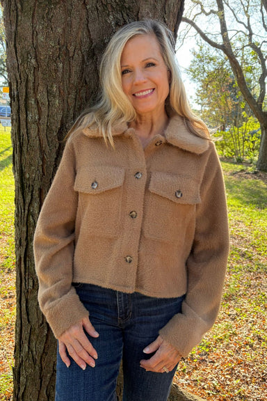 A smiling blonde woman wearing a cropped camel brown sherpa fleece jacket and dark blue jeans, standing outdoors next to a large tree trunk in a grassy park area.