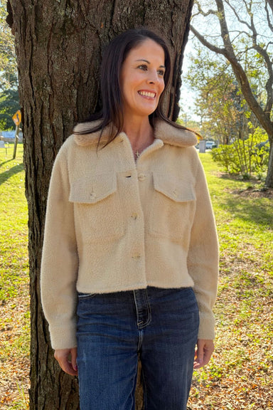 A woman wearing a beige fuzzy cropped jacket with a collar and flap chest pockets, paired with dark blue jeans, standing outdoors against a tree trunk in a grassy park setting.