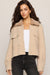 A woman modeling a cropped beige fuzzy jacket with a plush collar, worn over a white top and black jeans, against a light gray studio background.