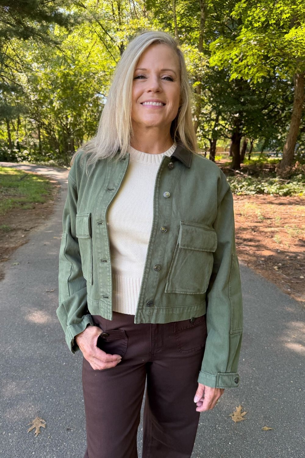 A woman standing on a paved path in a wooded outdoor setting, wearing an olive green cropped utility jacket over a cream ribbed sweater and dark brown pants.