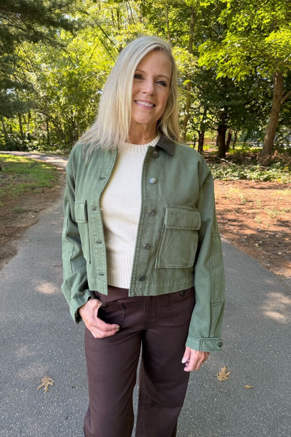 A woman standing on an outdoor paved path wearing an olive green cropped utility jacket over a cream ribbed top and dark brown pants.