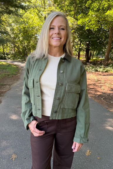 A woman standing on a paved path in a wooded outdoor setting, wearing an olive green cropped utility jacket over a cream ribbed sweater and dark brown pants.